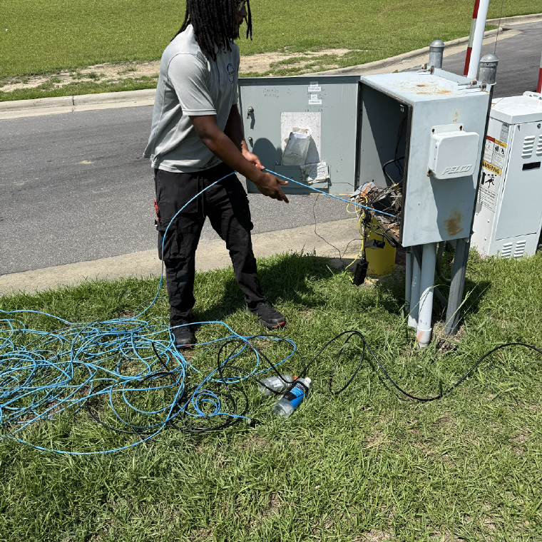 Technician pulling category cable to an outdoor equipment cabinet beside a gate arm